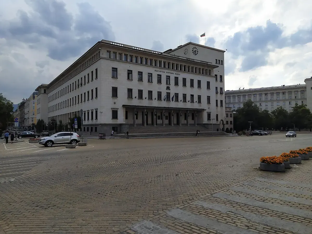 Сградата на Българската народна банка в София с часовник и знамена, пред нея павиран площад с паркирани коли